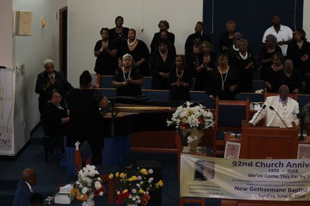 Choir on church stage near pulpit, with white flowers and a banner celebrating church anniversary.