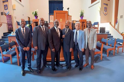 Six men in suits posing on a church stage with blue carpet and wooden chairs behind them