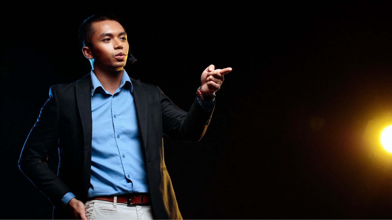 A presenter speaks to an audience in a bright room, gesturing with both hands while standing by a white podium.