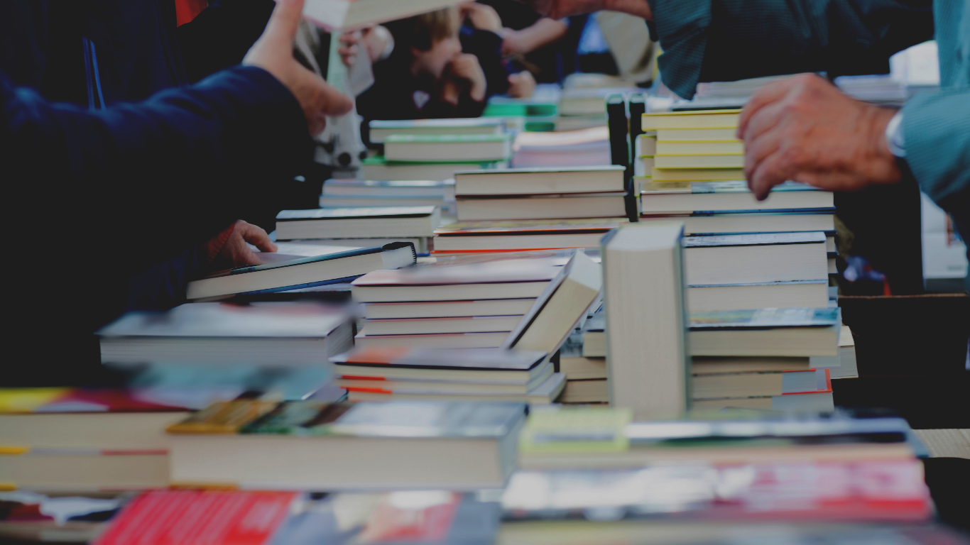 A hand holding a black pen signs an open book on a wooden surface as another hand reaches out toward it.