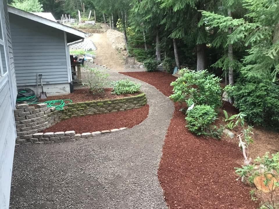 Mulch-covered garden path curving past a gray house and green shrubs in a wooded yard