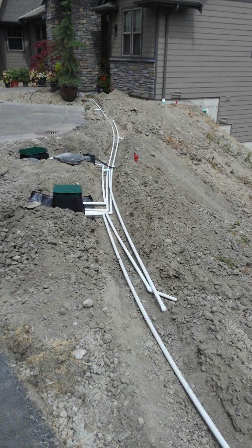 Trenched construction site beside a house with white PVC pipes laid along a dirt slope.