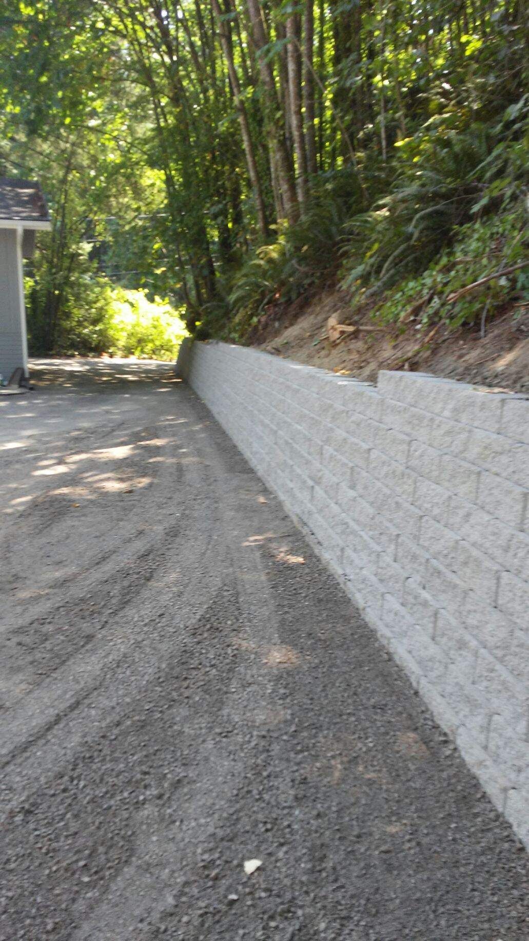 Gravel driveway beside a gray retaining wall on a wooded hillside, with a building edge at left.