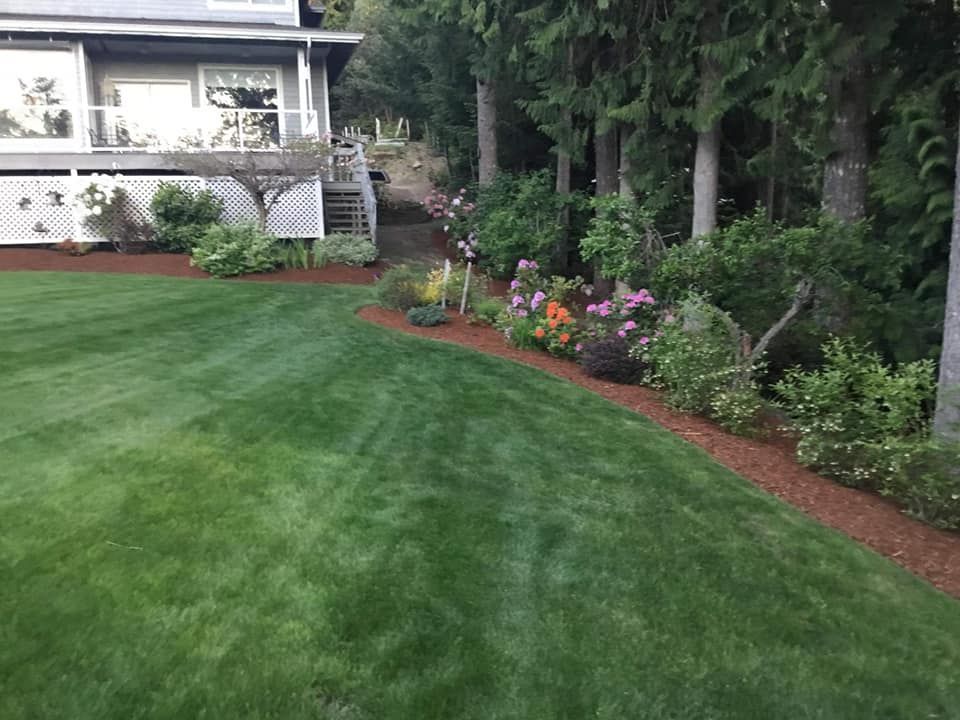A landscaped backyard with a green lawn, flower beds, and a house patio at the edge of a wooded area.