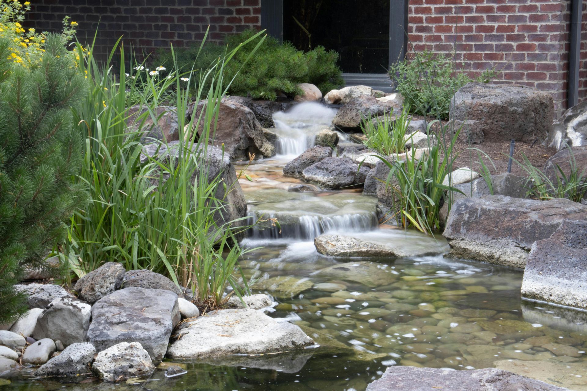 Small garden waterfall cascading over rocks into a pond beside brick walls and green plants