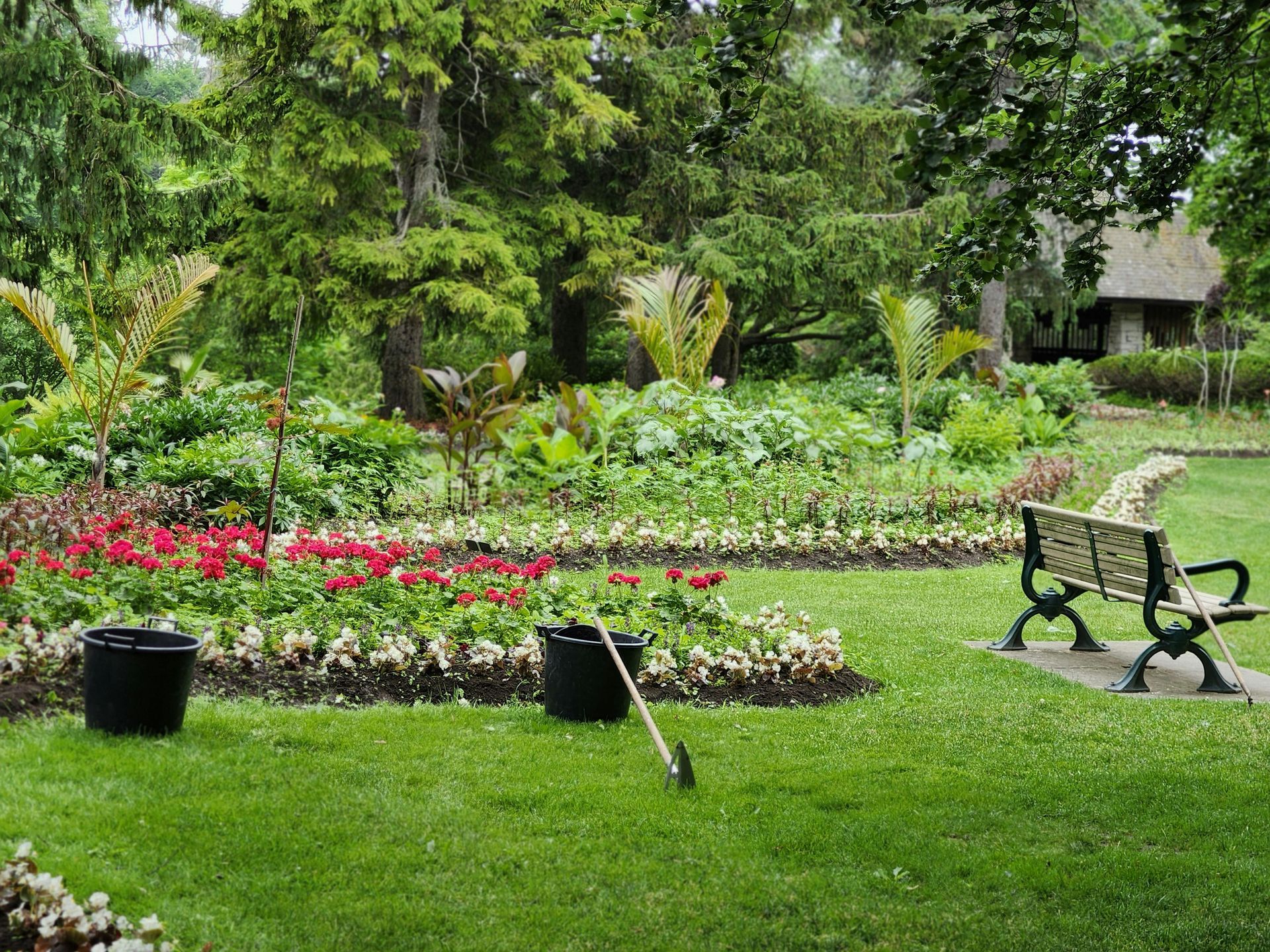 Garden with colorful flower beds, green lawn, trees, and a bench beside a small path.