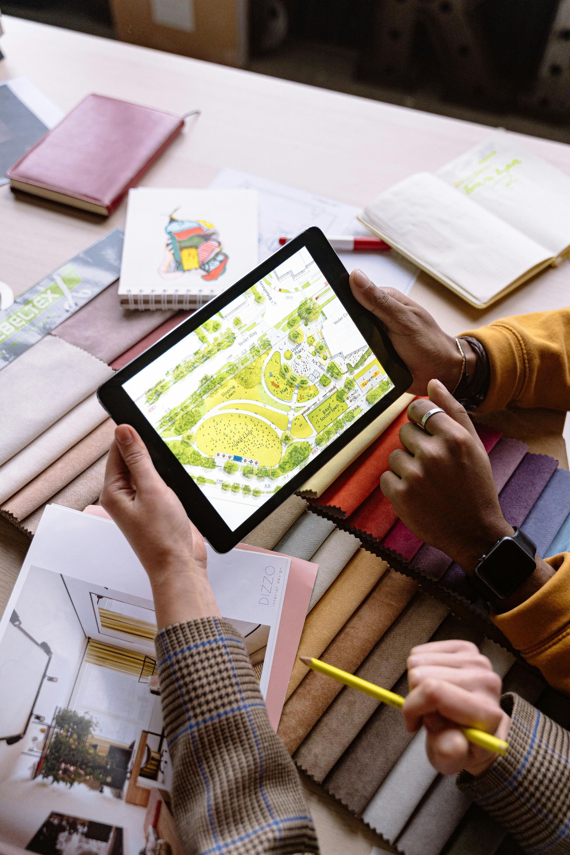 Hands using a tablet with a colorful map, surrounded by notebooks and pens on a desk.