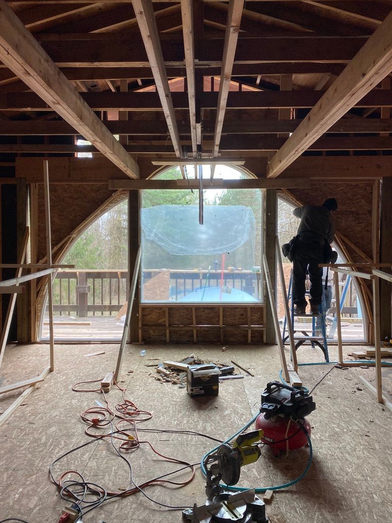 Construction site interior with exposed beams, tools on the floor, and a large window overlooking trees and a building