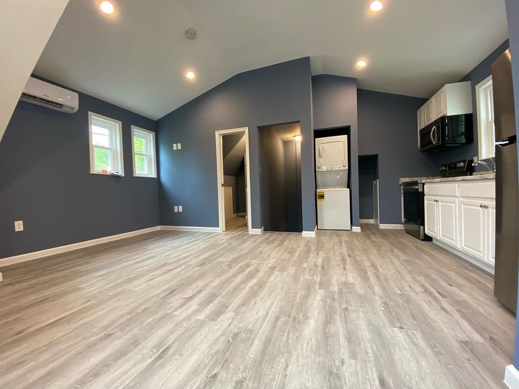 Empty modern living room with dark walls, light wood floors, and a small kitchen area.