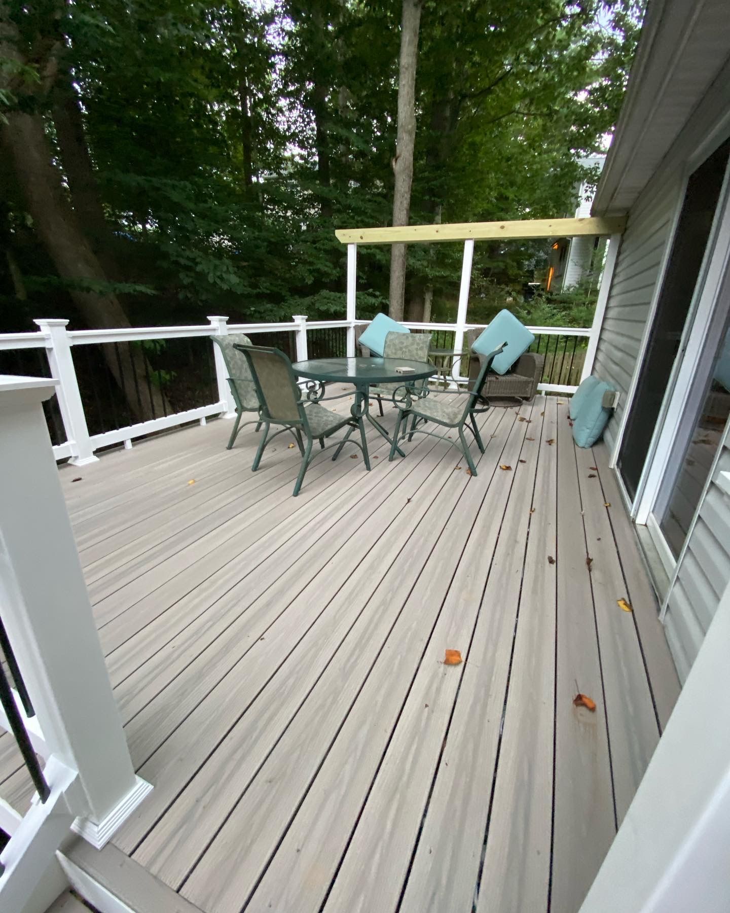 Outdoor deck with glass table, chairs, and turquoise cushions beside a house and trees