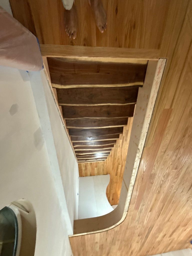 Narrow wooden staircase with white walls, viewed from above toward a bright lower floor