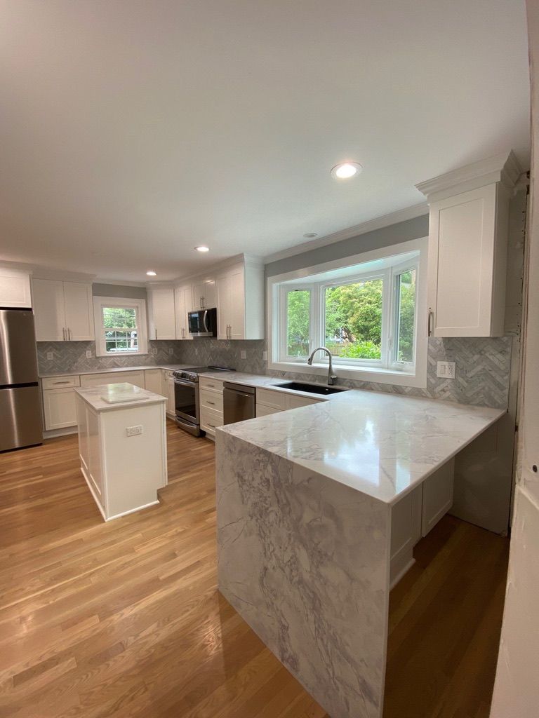Bright modern kitchen with white cabinets, marble island, wood floors, and large windows overlooking greenery