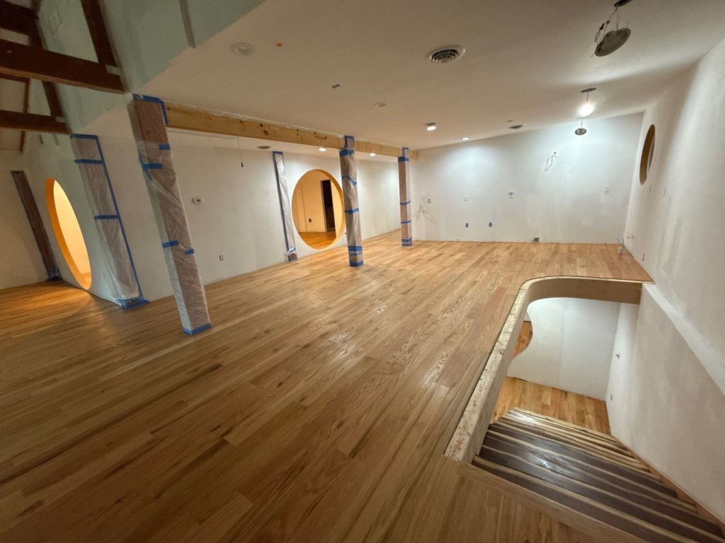 Empty loft room with wood floors, white walls, exposed beams, and a stair opening in the foreground