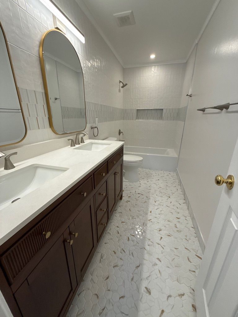 Bright bathroom with double vanity, large mirror, and bathtub along a narrow tiled floor.