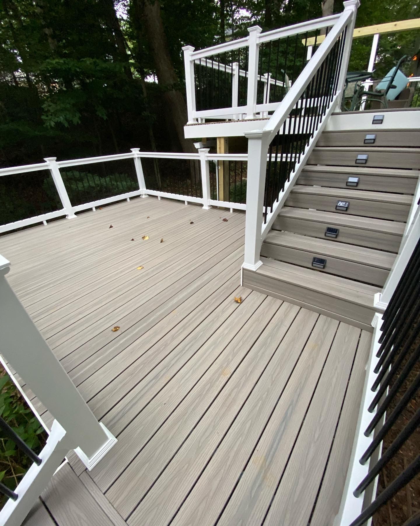 Wooden deck with white railings and stairs leading to an upper landing, surrounded by trees.