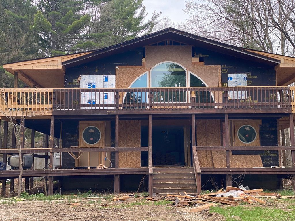 Two-story cabin under construction with wood deck, large arched window, and exposed framing amid trees.