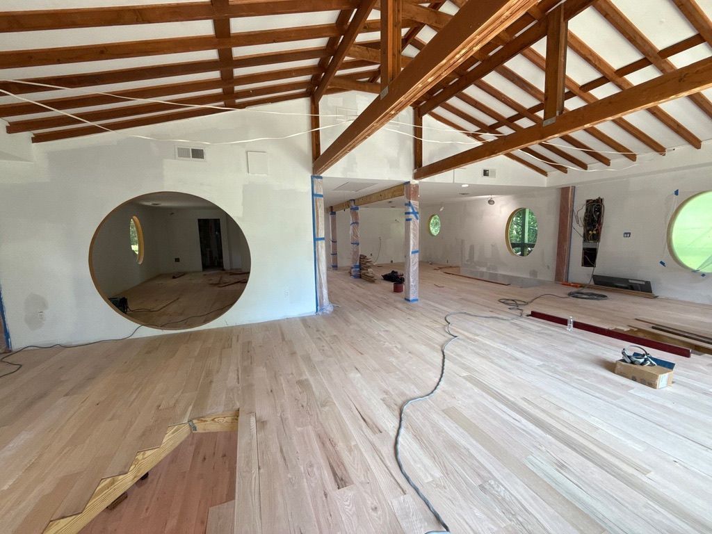 Spacious unfinished room with exposed wooden beams, round windows, and wood flooring under construction