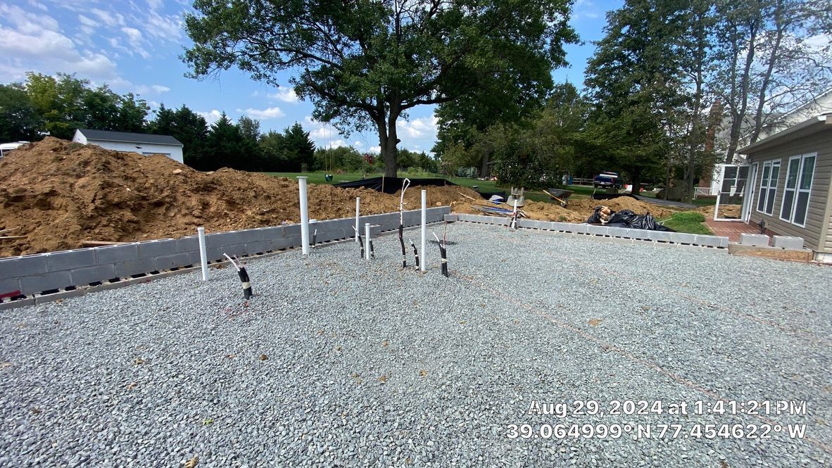Gravel-covered construction site with exposed white pipes, dirt piles, and trees beside a house