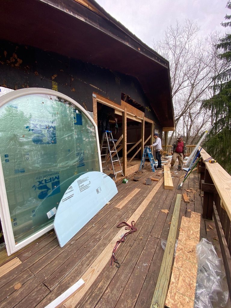House exterior under renovation with workers, exposed framing, and windows removed on a wooden deck