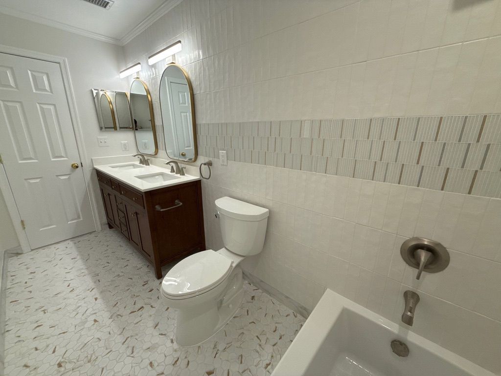Bright bathroom with white tile, wooden vanity, toilet, and bathtub.