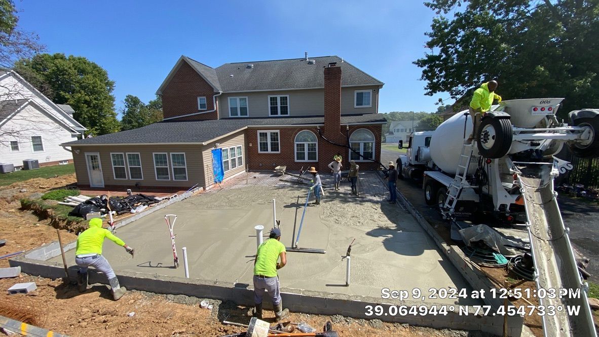 Construction crew pouring concrete for a new patio beside a brick house, with a cement mixer truck nearby