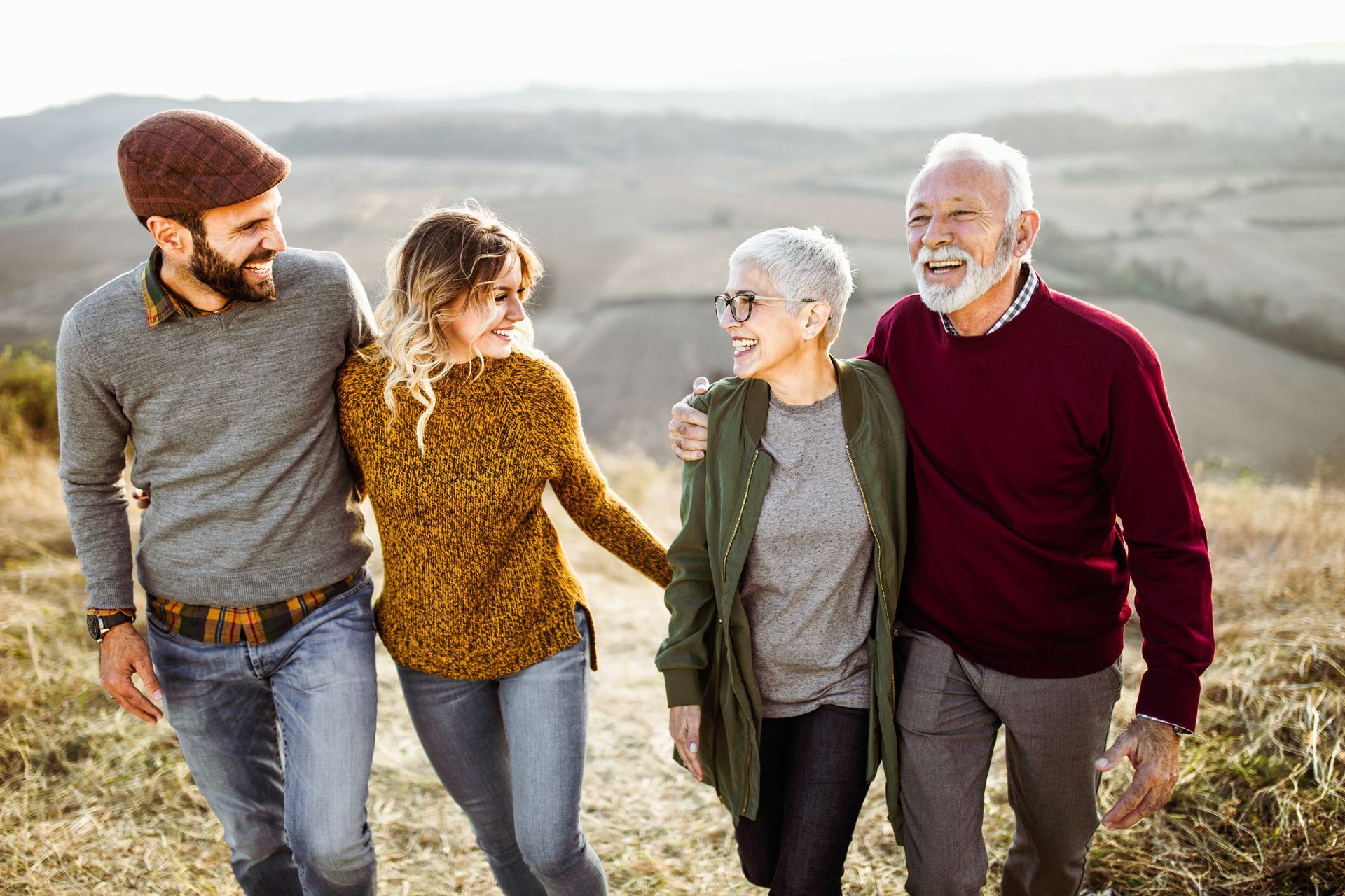 Four friends walking together outdoors on a sunny hillside, smiling and chatting.