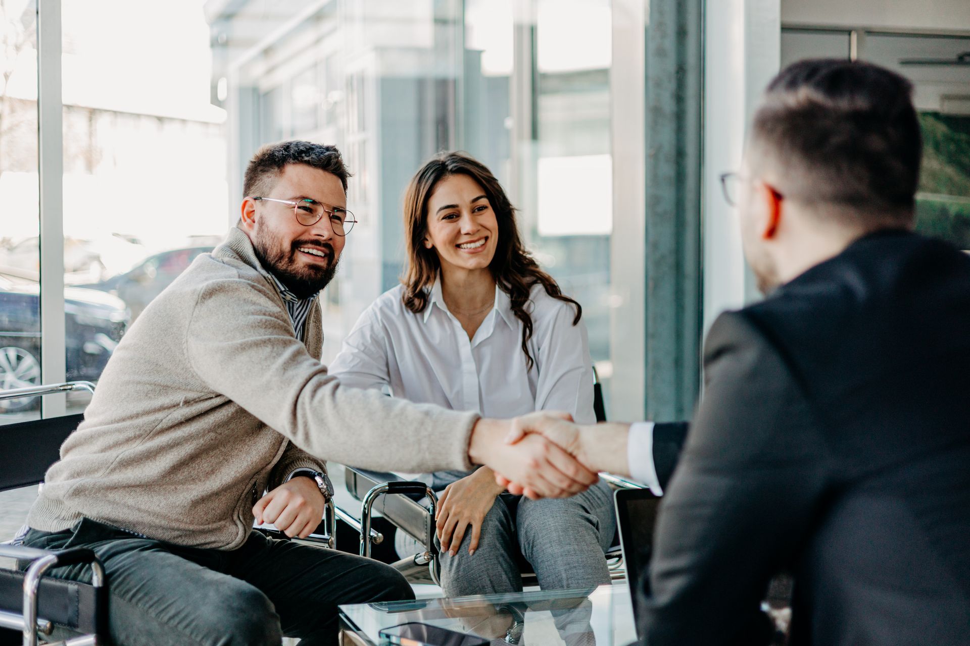 Two people shaking hands in a bright office, with a third person seated nearby.