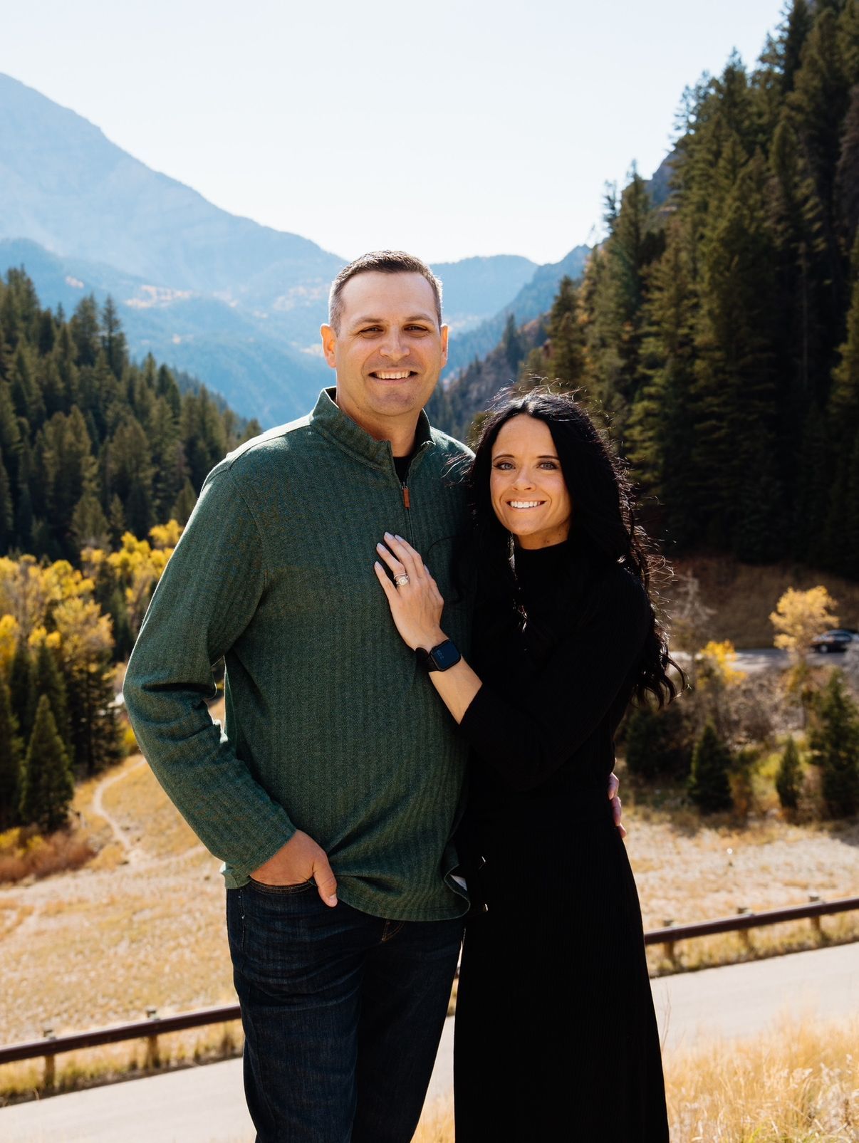 Couple standing together in a mountain valley, smiling, with evergreen trees and hills behind them
