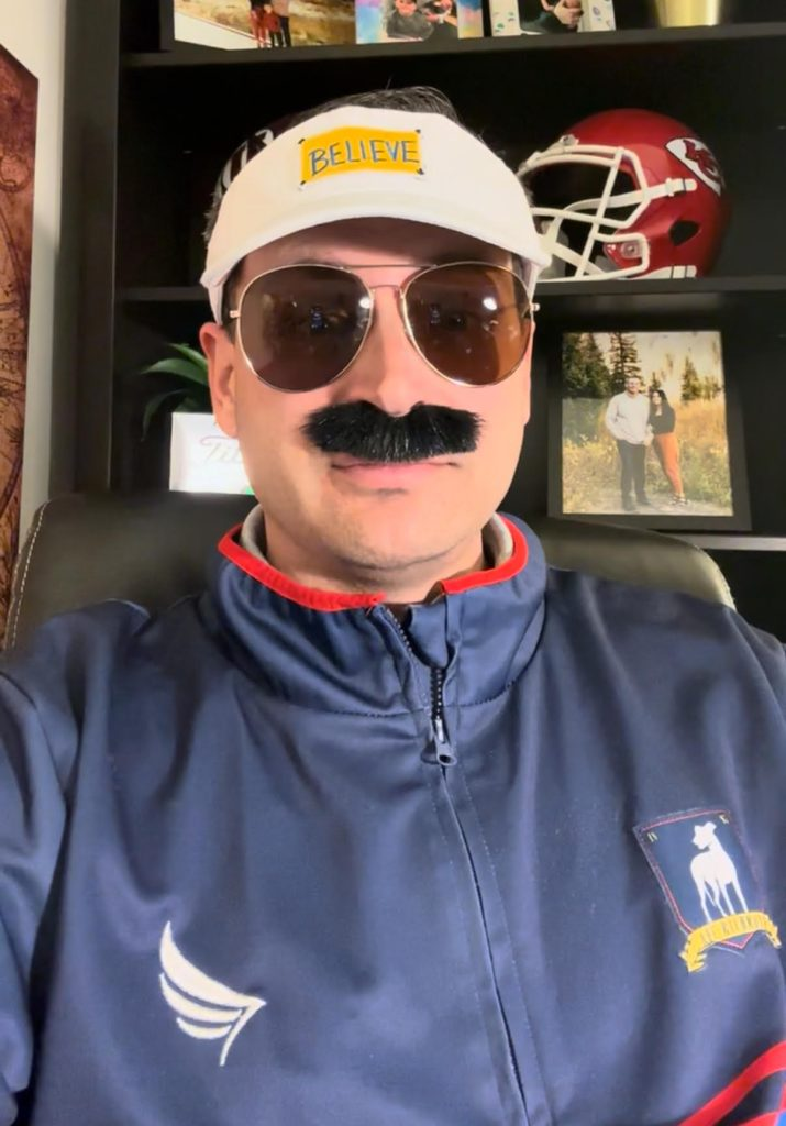 Man wearing sunglasses, fake mustache, and cap, posing indoors with sports memorabilia behind him