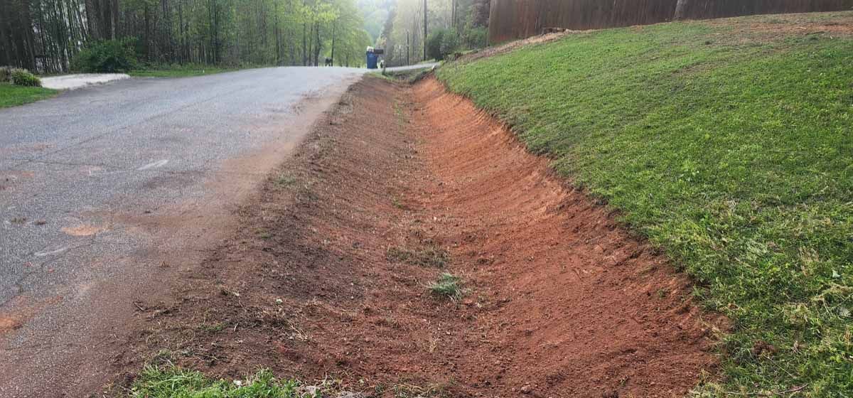 A roadside ditch with reddish soil, next to a paved road and grassy bank.