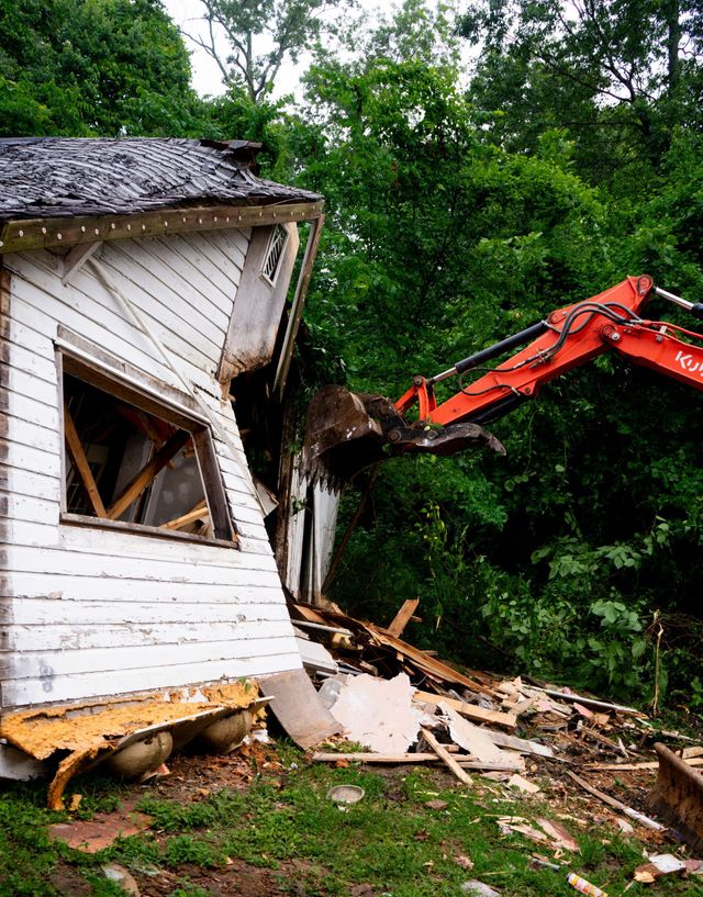 Excavator demolishes a white building; orange arm, green foliage, debris on grass.