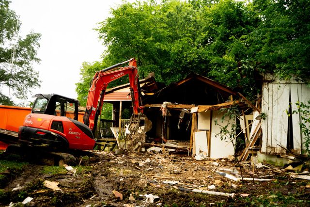 A red excavator demolishes a white house with debris scattered on the ground. Trees in the background.