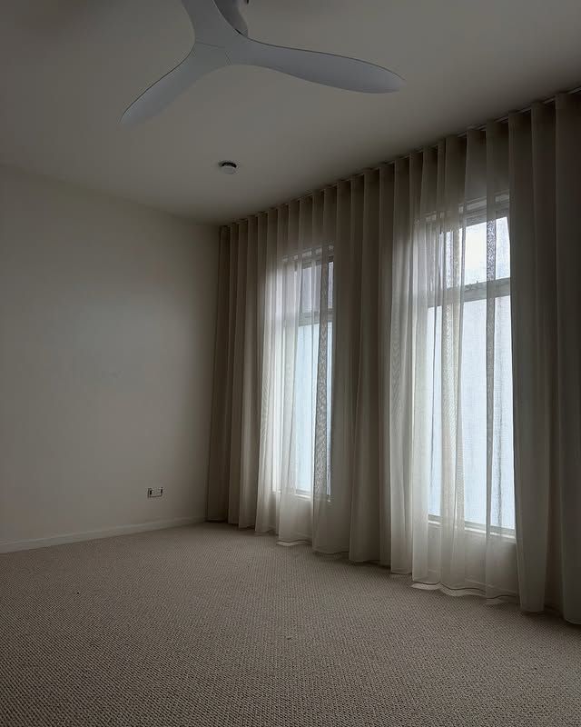 Empty room with neutral-colored carpet, walls, and sheer curtains covering two windows. A ceiling fan is mounted.