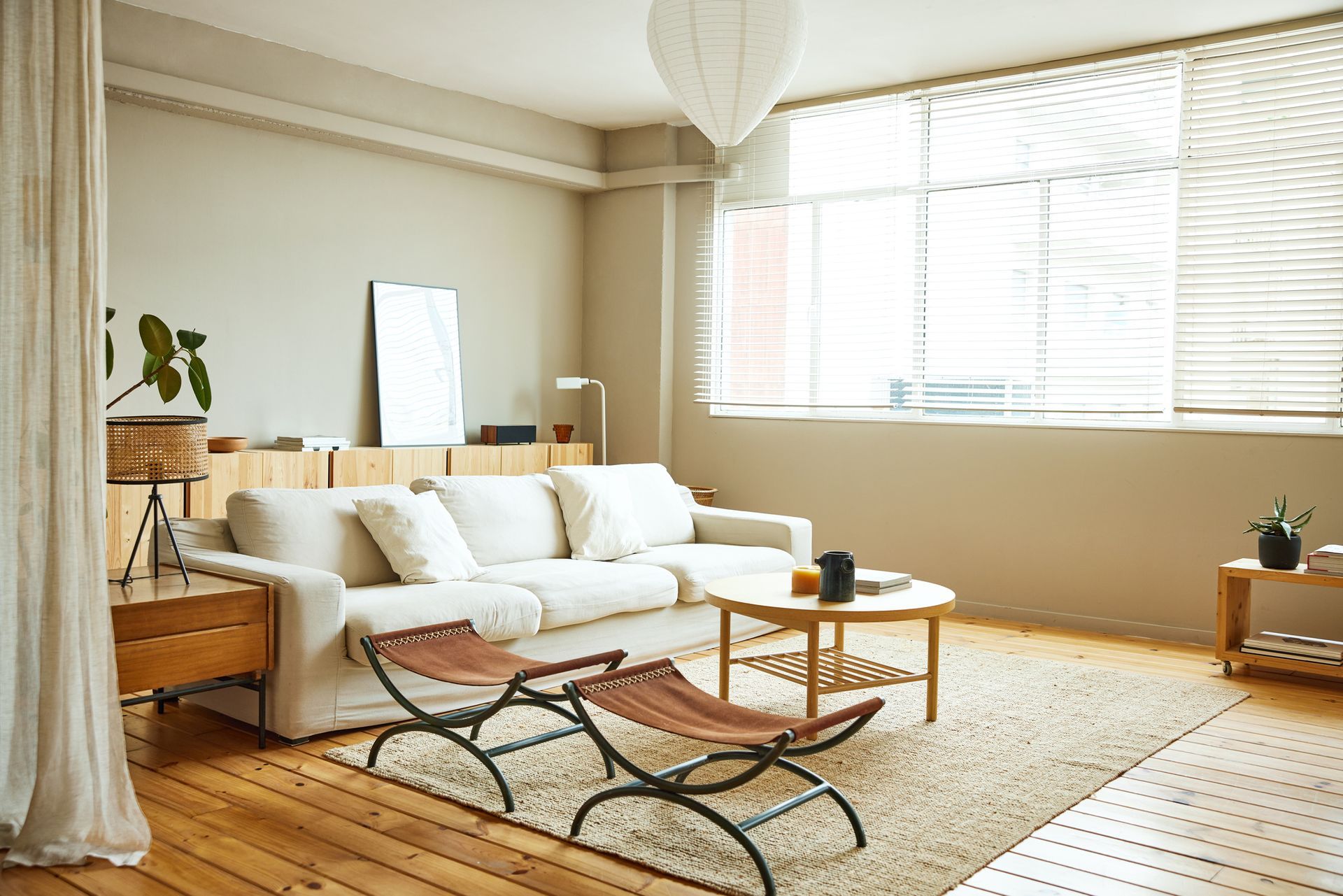 A bright living room with a white sofa, wooden coffee table, and two brown leather stools. Large window with blinds.