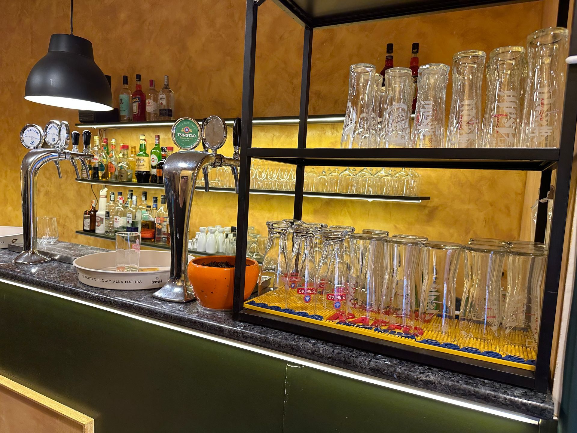 Bar con file di bicchieri, spillatore di birra e bottiglie sugli scaffali, illuminato da luci calde