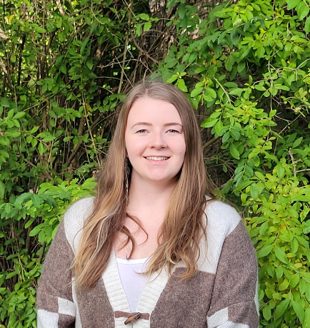 A woman in a brown and white sweater is standing in front of a bush.