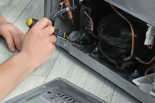 Unas manos usan un destornillador para reparar el interior de un pequeño electrodoméstico con cables y compresor a la vista.