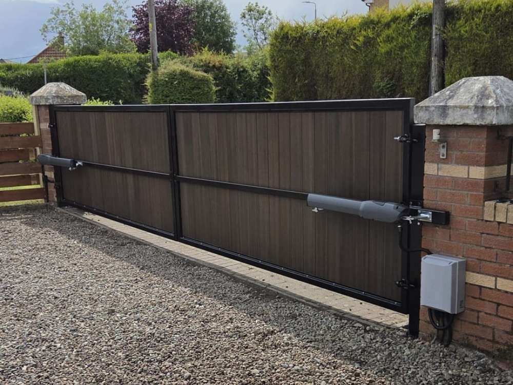 Composite wood-effect electric swing gates in a black frame between brick pillars on a gravel driveway