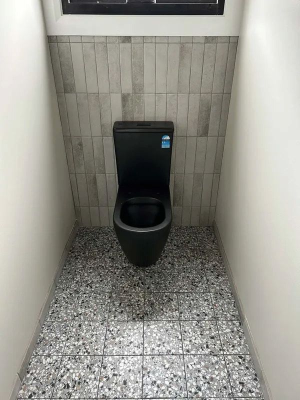 Black Toilet in a Small Tiled Restroom Stall With a Speckled Floor — The Plumbing & Drainage Co. in Campbelltown, NSW