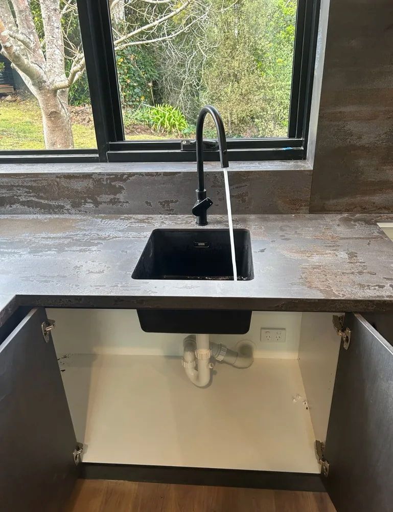 Kitchen Sink With Black Faucet Beneath a Window, With Open Cabinet — The Plumbing & Drainage Co. in Mittagong, NSW