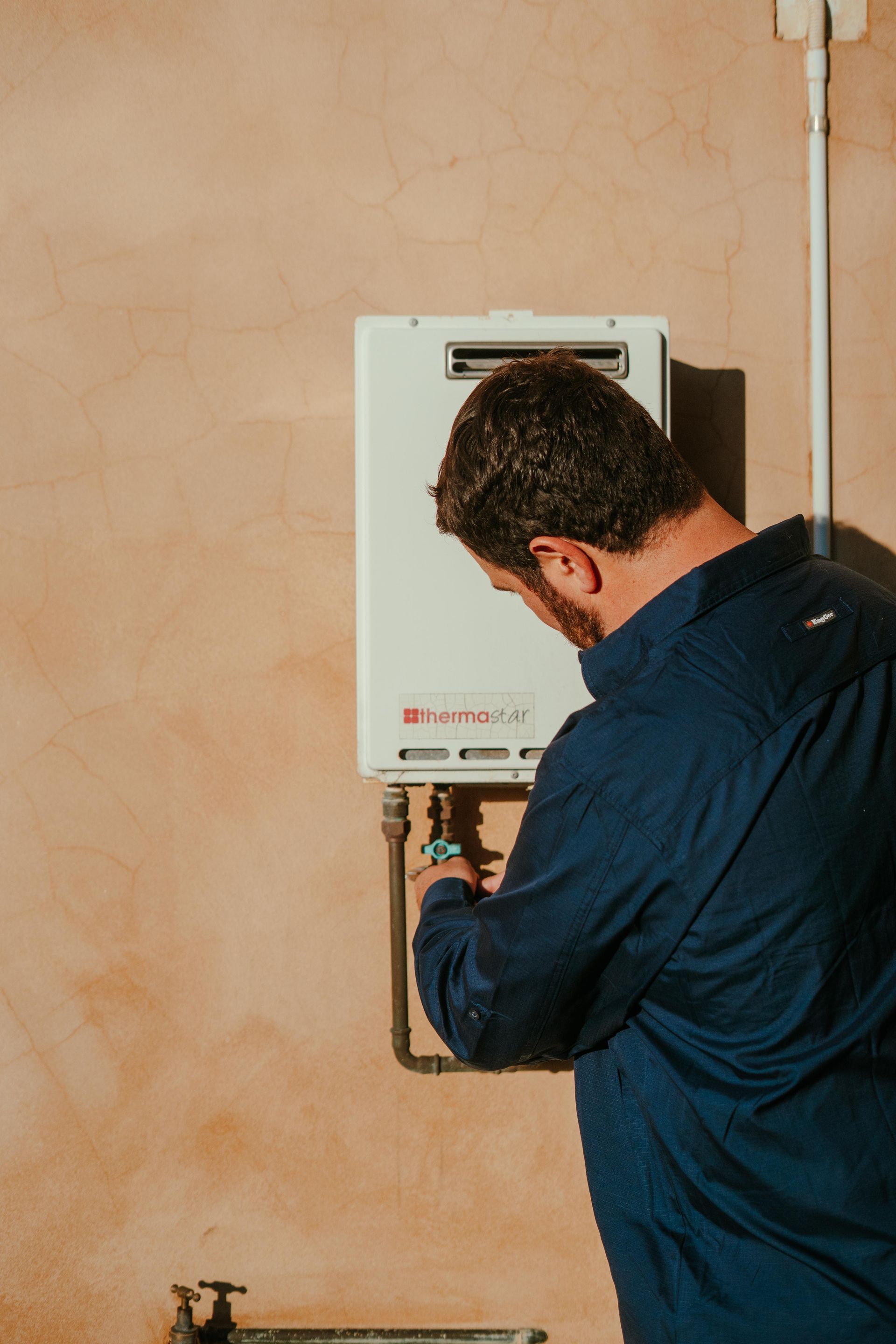Technician inspecting a wall-mounted boiler, viewed from behind in a beige room — The Plumbing & Drainage Co. in Shellharbour, NSW