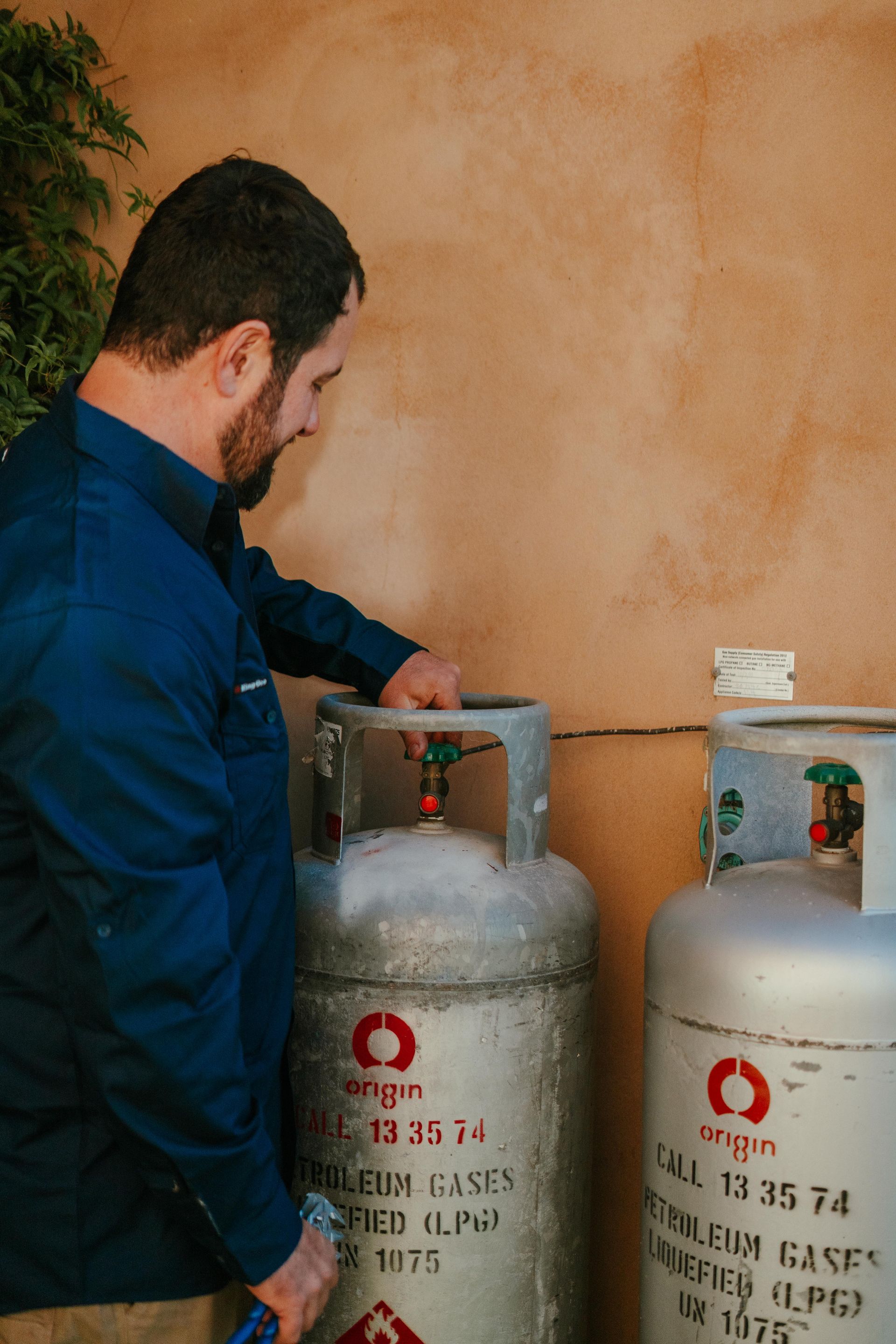 Man in blue shirt checking two silver gas cylinders against a wall. — The Plumbing & Drainage Co. in Southern Highlands, NSW