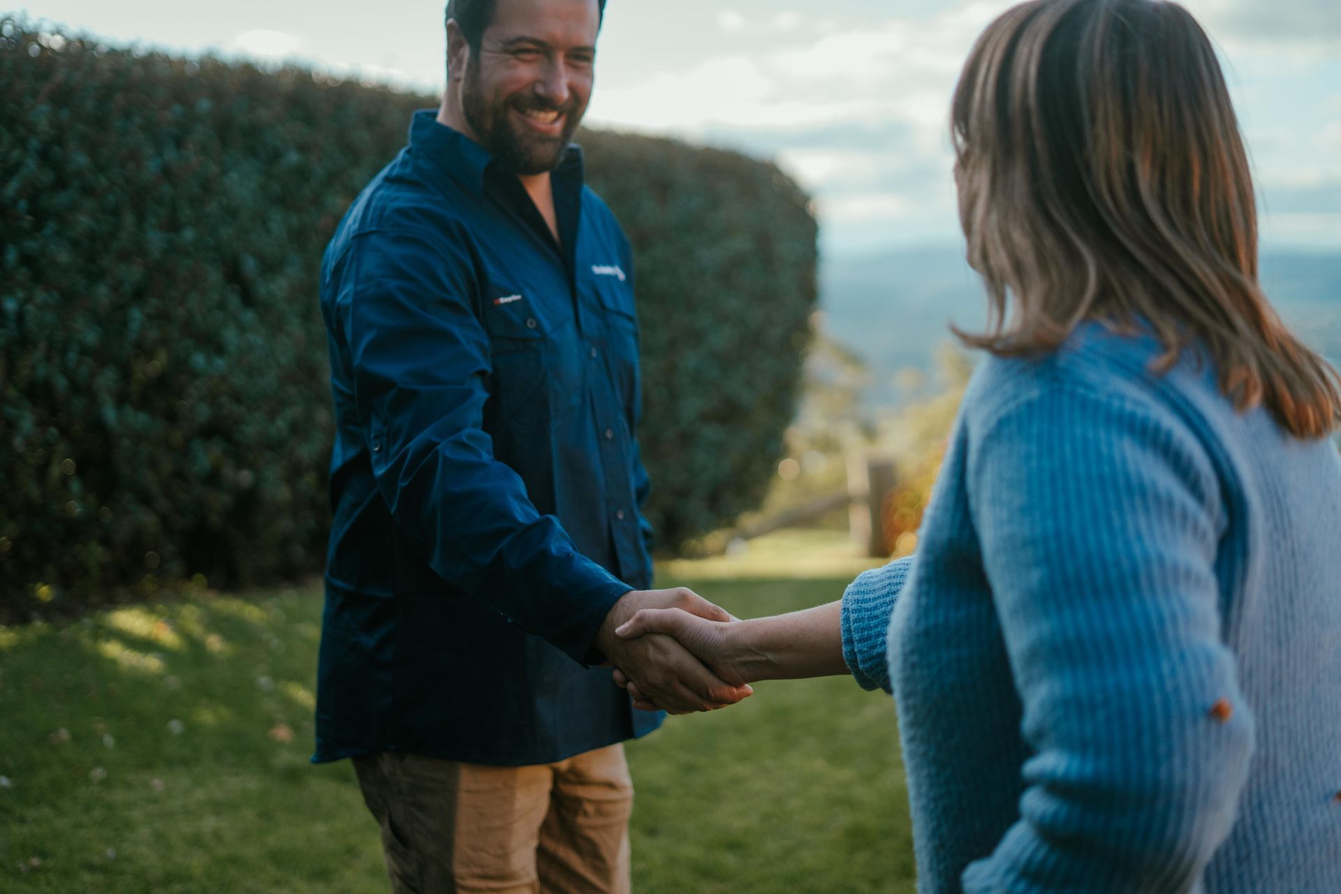 Two people shaking hands outdoors beside a hedge, one in a blue shirt and the other in a light blue sweater. — The Plumbing & Drainage Co. in Mittagong, NSW