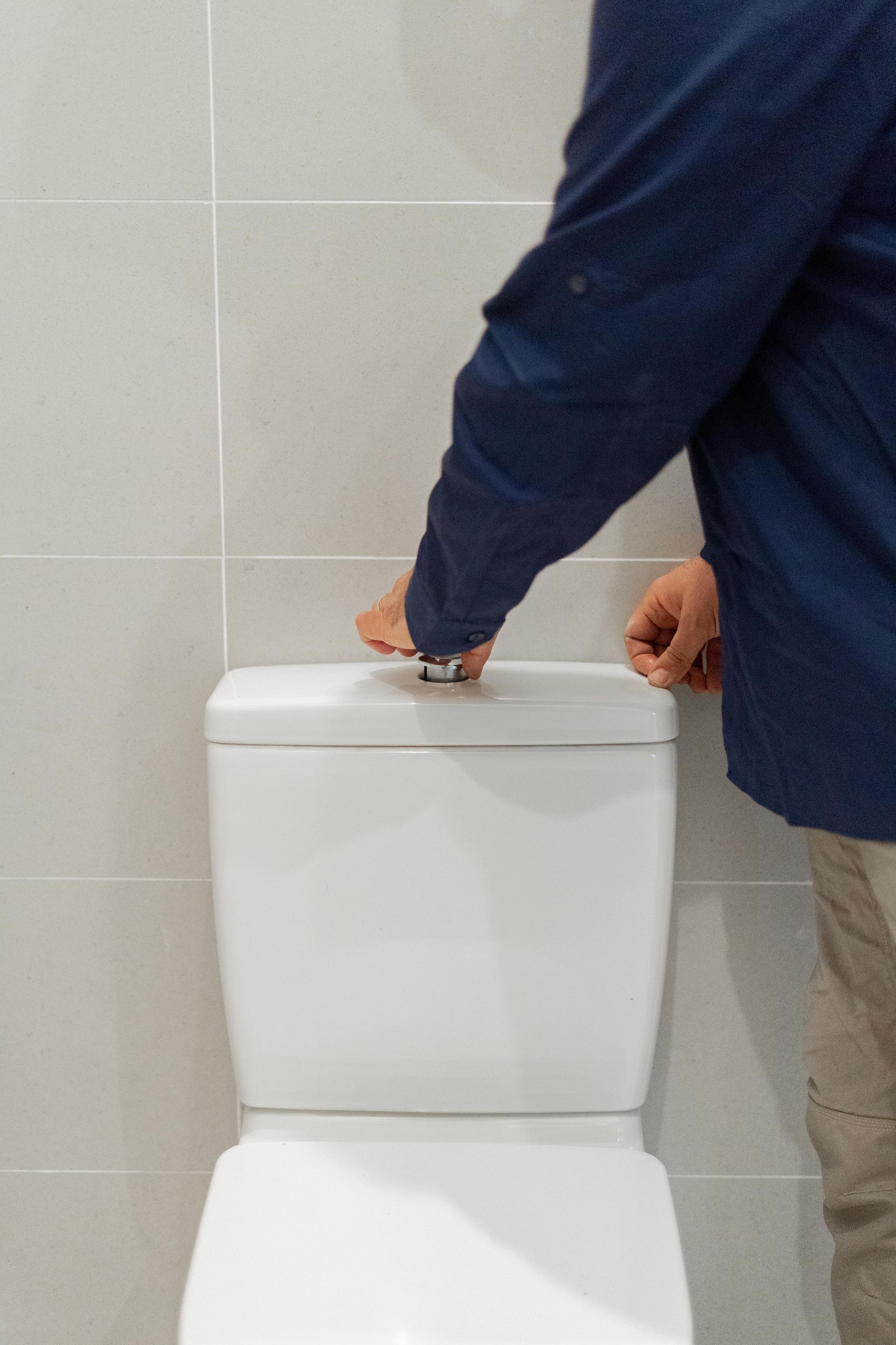 Person in a blue jacket opening a white toilet tank lid in a tiled restroom. — The Plumbing & Drainage Co. in Mittagong, NSW