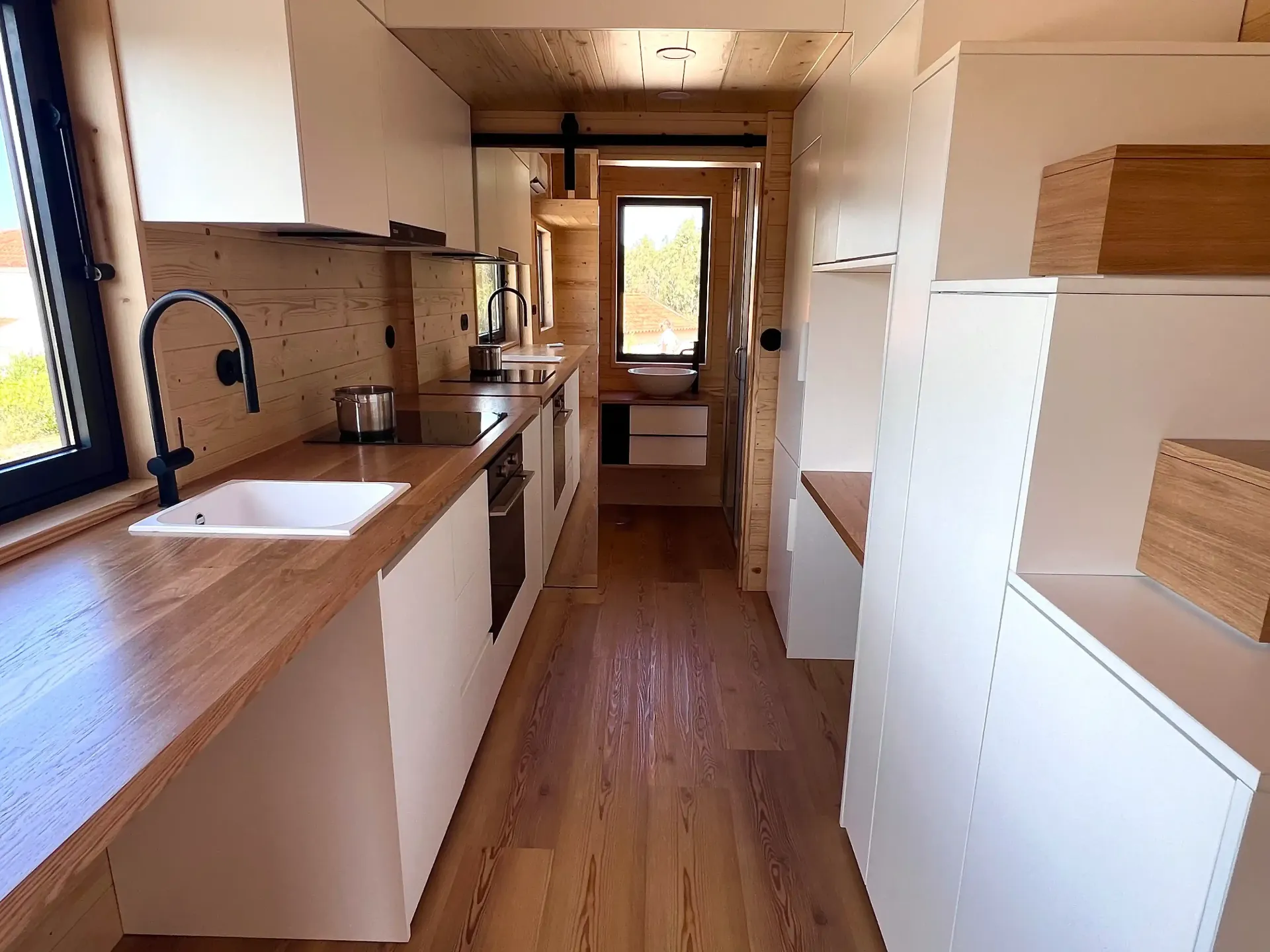 Narrow, bright interior of a tiny home with kitchen, cabinets, and wood paneling.