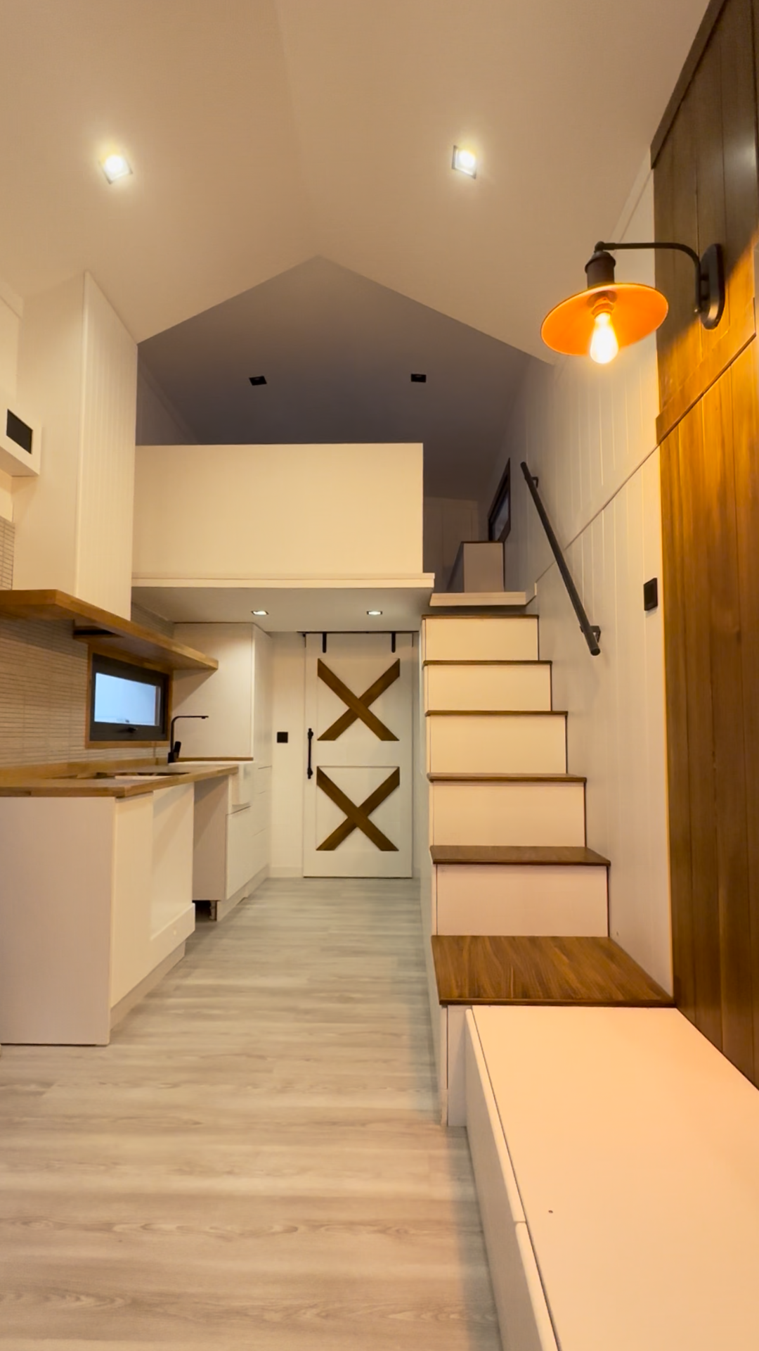 Interior of a tiny home with loft and staircase, light wood, white walls, and an orange light fixture.