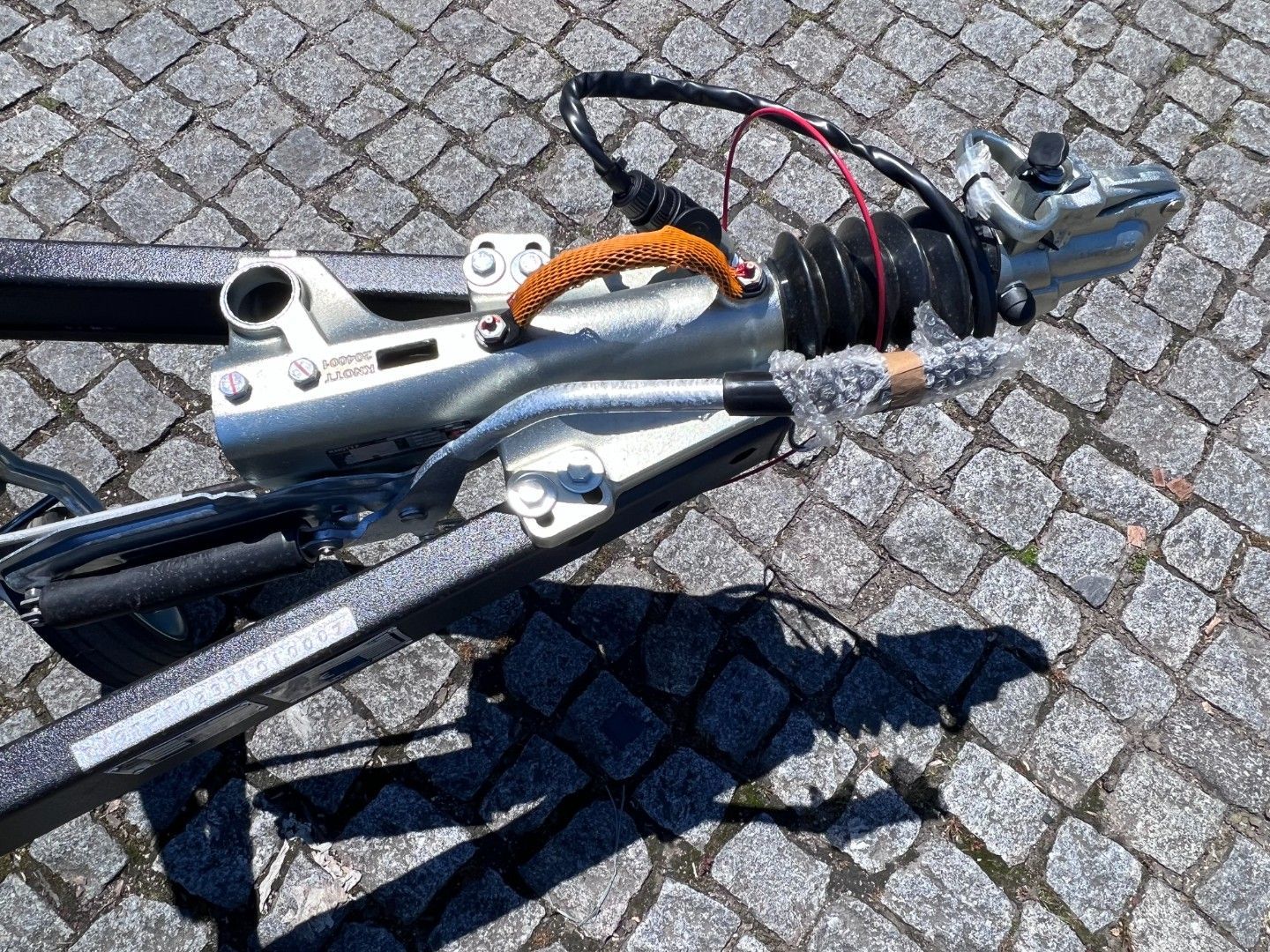 Silver trailer hitch on a gray frame with a black cable and orange handle on cobblestone.