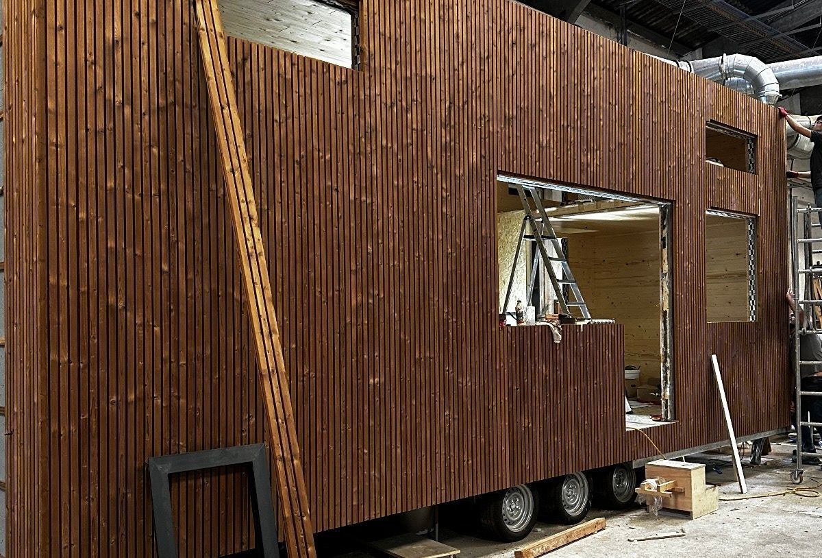 Wooden tiny house exterior under construction. Brown vertical siding, window frames, and wheels are visible.