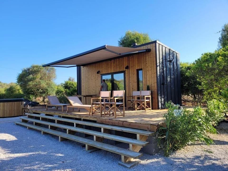 Wooden cabin with a deck, awning, and chairs in a gravel yard.
