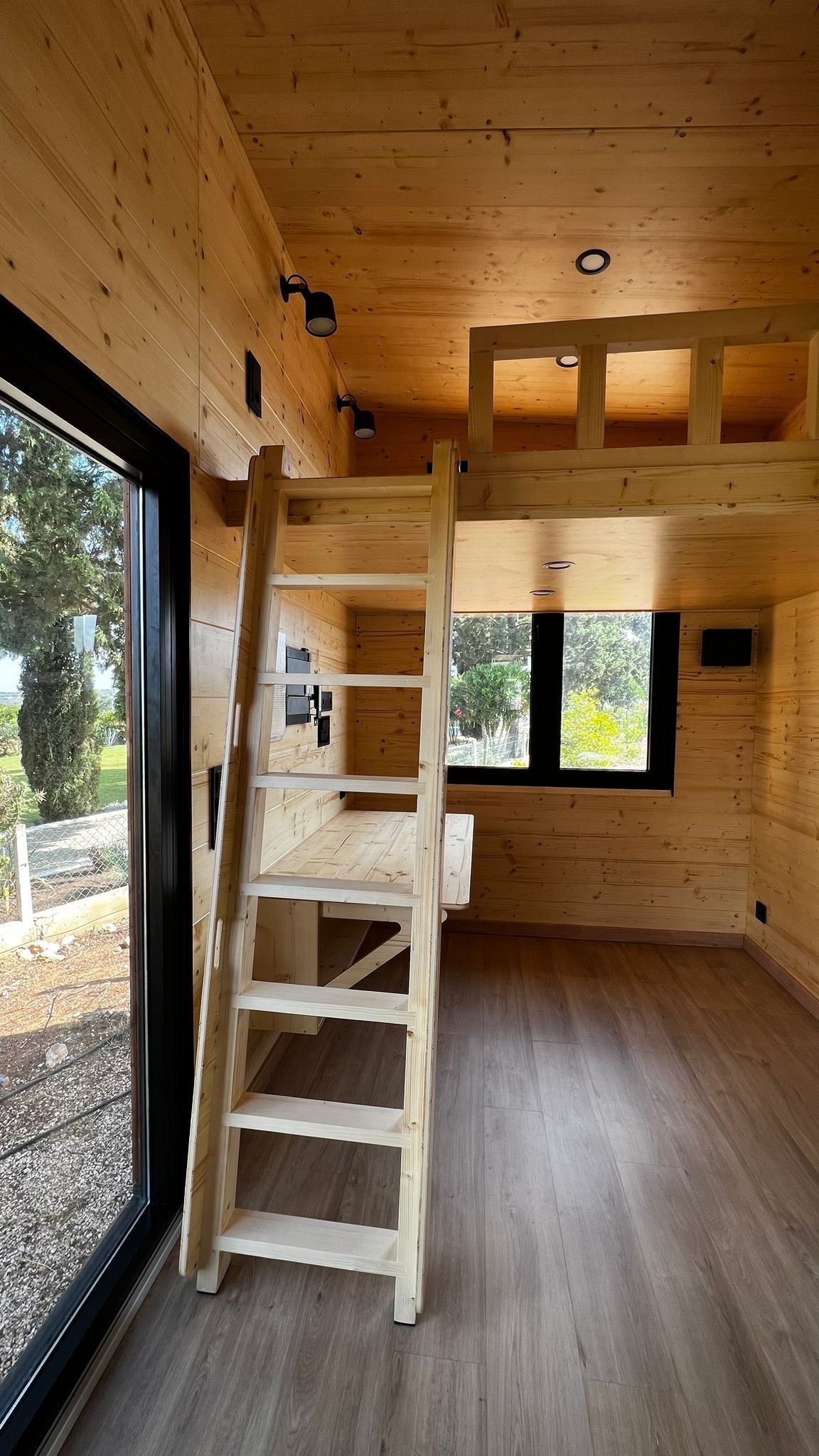 Interior of a tiny house with a wooden ladder to a loft, large window, wood paneling, and a desk.